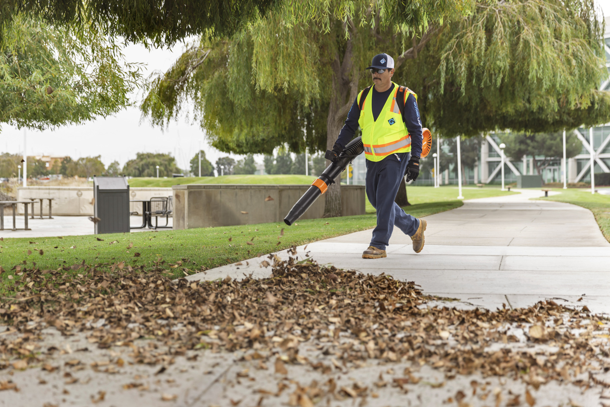A man wearing work gloves is using a STIHL leaf blower to blow leaves off the sidewalk