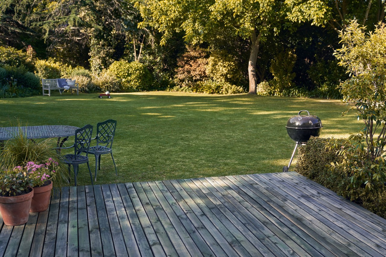 Terrasse en bois devant un jardin verdoyant avec barbecue, chaises et pots de fleurs