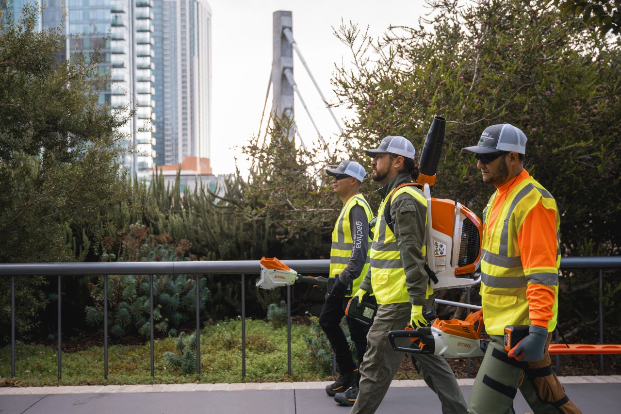Drie hoveniers lopen met werkkleding en STIHL machines in een stadsomgeving.